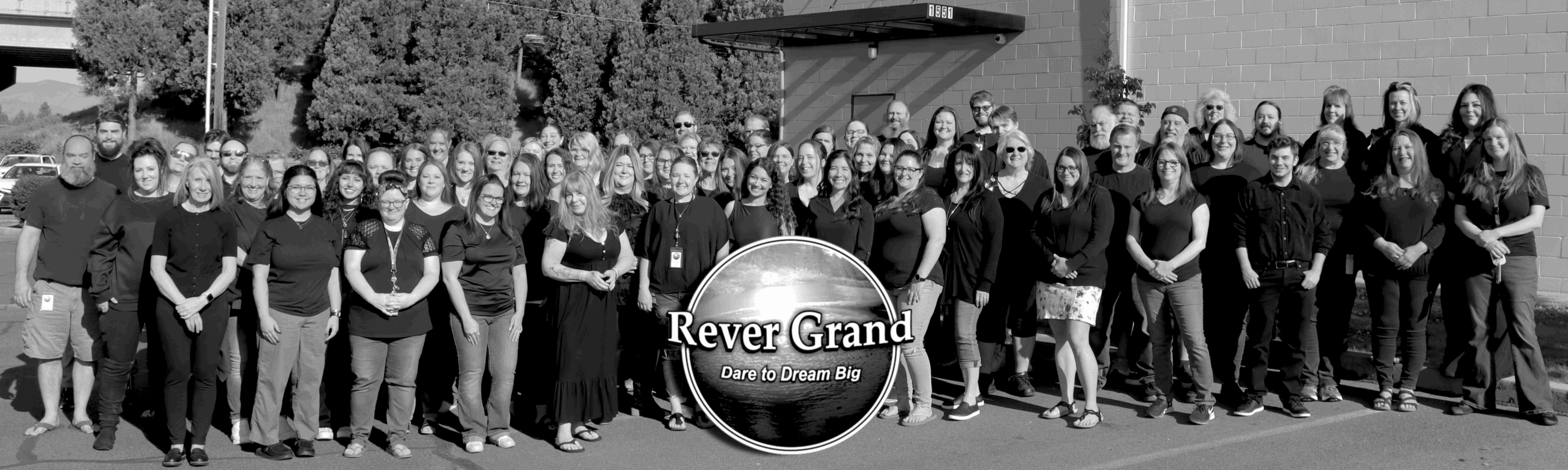 A black and white photo of the Rever Grand admin staff, standing outside of the corporate office in a team photo. The Rever Grand logo is positioned in the center in front of the team members.